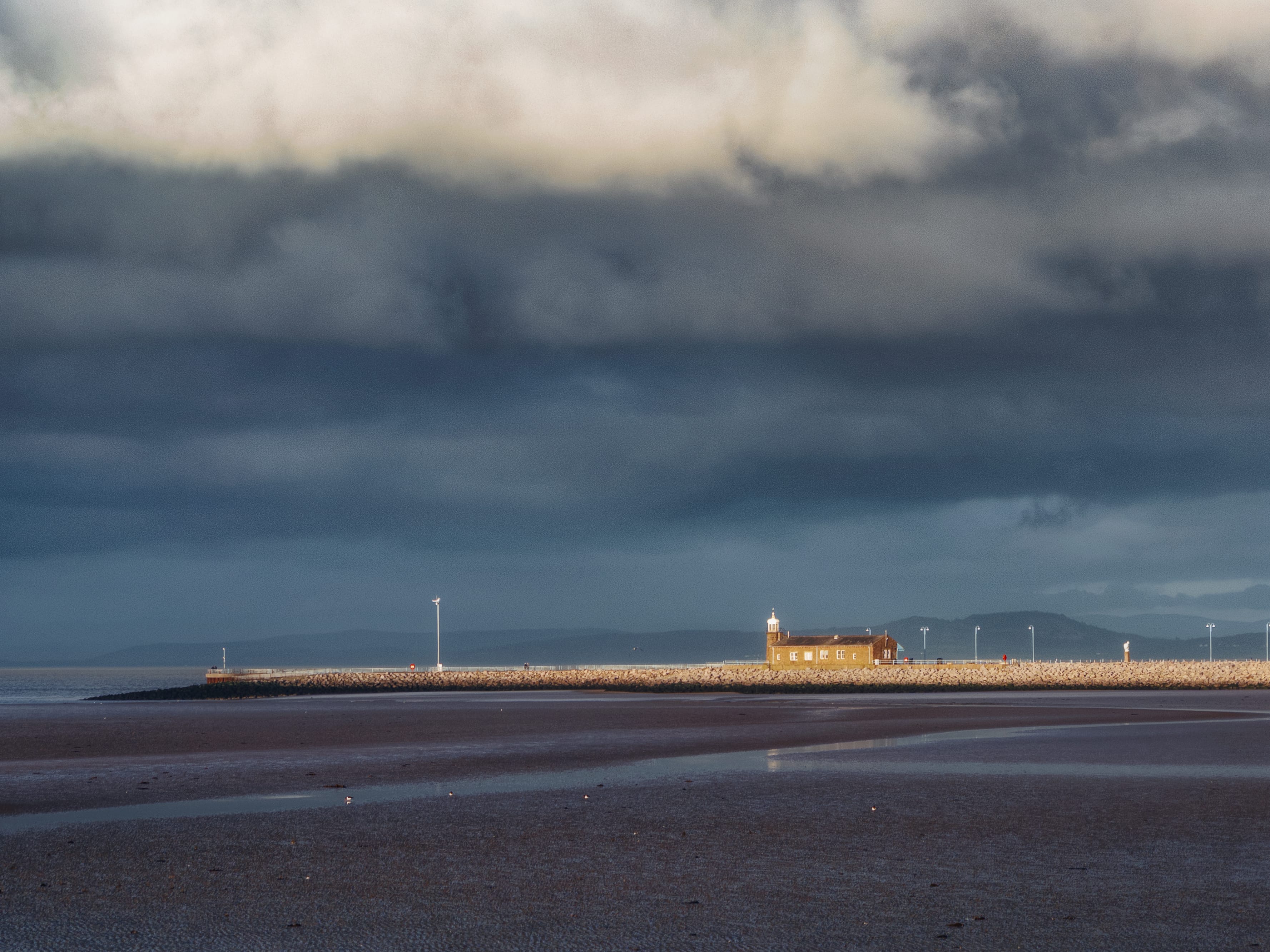 A dramatic view of the Stone Jetty at Morecambe, captured under a heavy, brooding sky filled with dark grey storm clouds. A narrow shaft of brilliant sunlight breaks through the gloom, spotlighting the long stone breakwater and its distinctive golden-stone building, which features a small white lighthouse tower. In the foreground, the vast expanse of Morecambe Bay's wet sands at low tide creates shimmering reflections of the moody sky, while the faint, hazy silhouettes of the Lake District fells are visible across the water on the horizon. The stark contrast between the luminous jetty and the deep shadows of the encroaching storm provides a powerful sense of atmosphere and scale along the Lancashire coast.