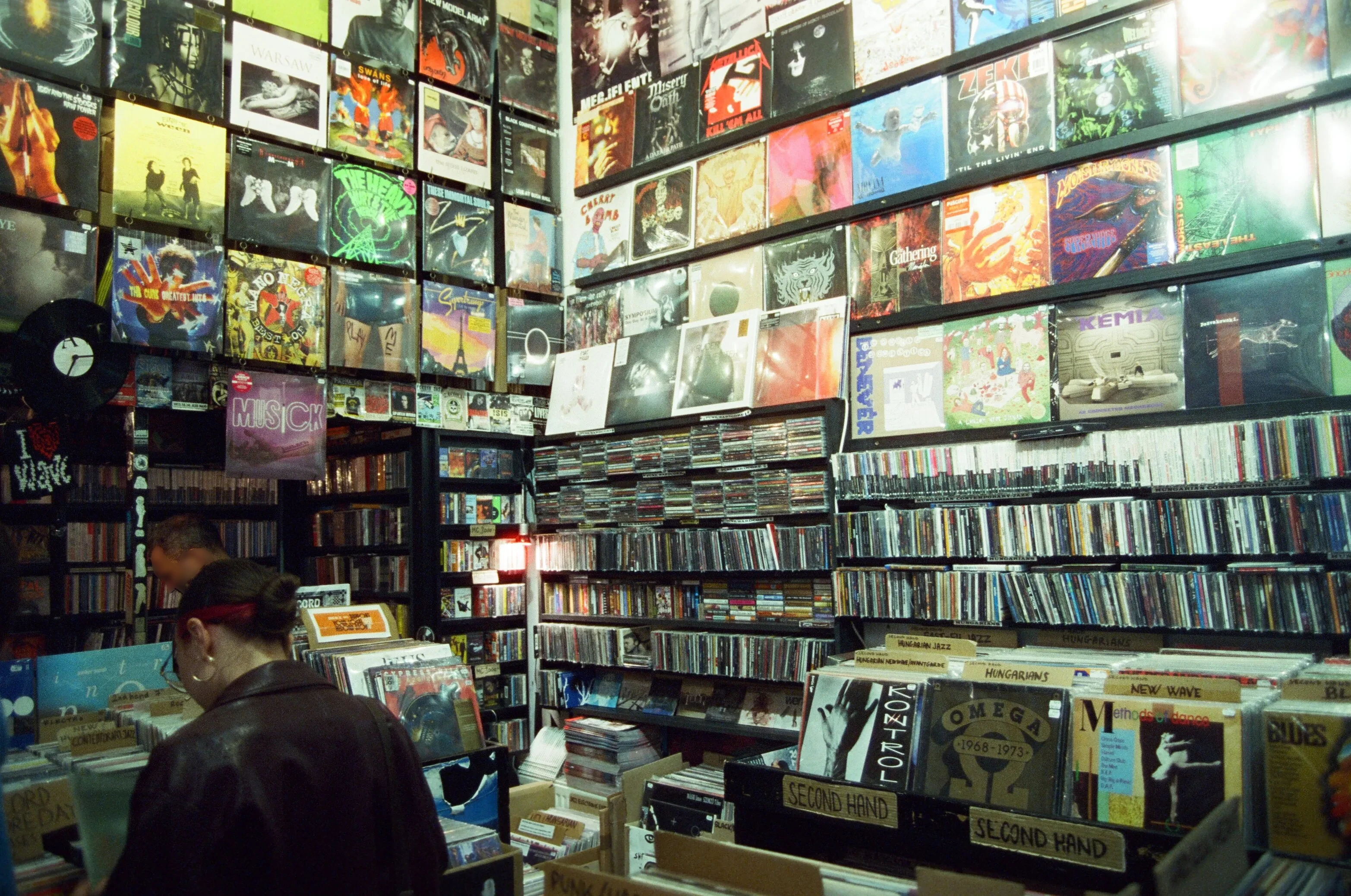 A film photograph inside a Budapest record store: album covers are displayed across tall walls, creating a colorful collage of music artwork. Shelves filled with CDs stretch along the room, while record bins labeled by genre and section fill the floor. A person in a dark jacket and a red streak in their hair browses through vinyl records near the front of the shop.