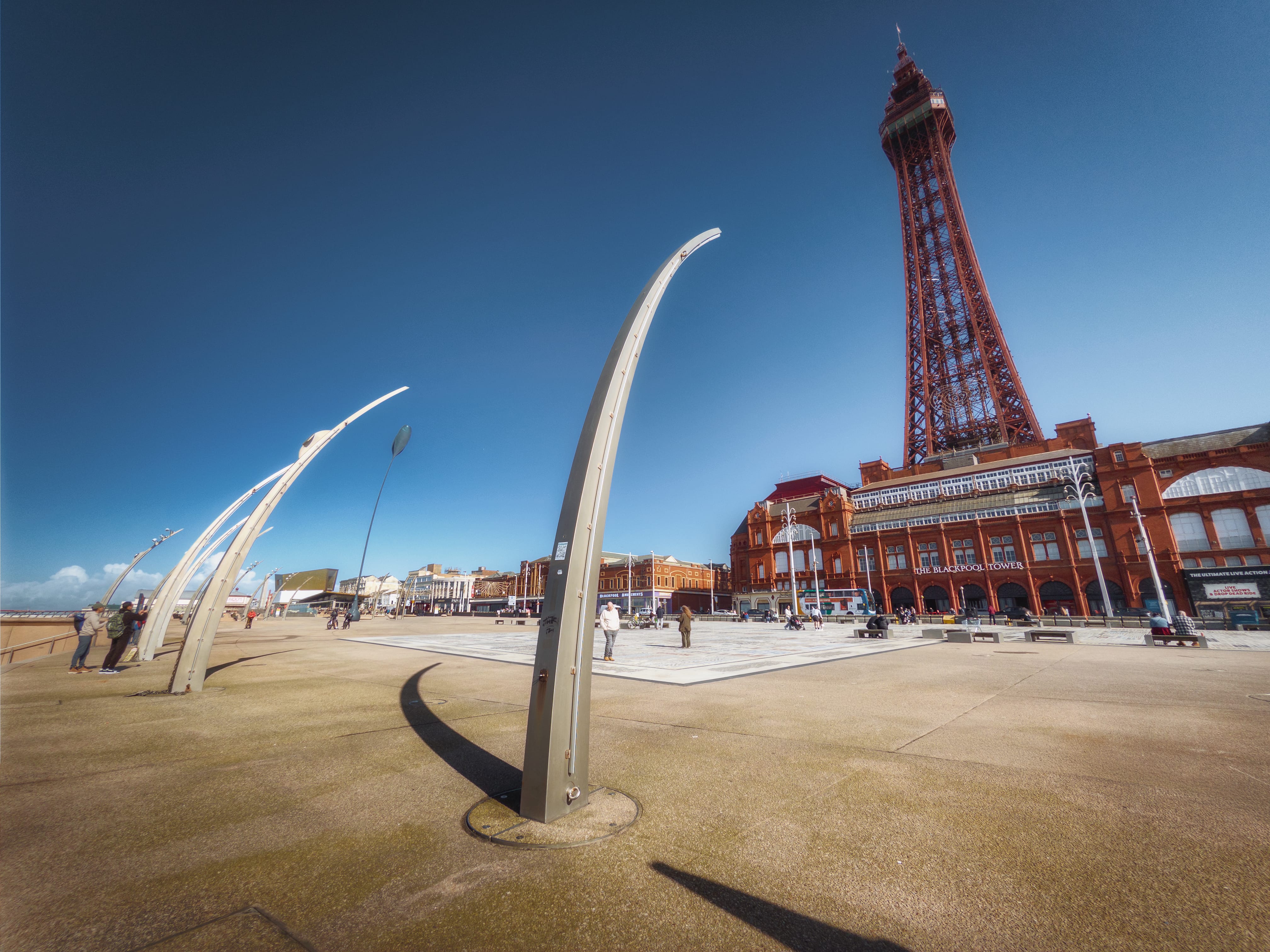 A seaside promenade at Blackpool featuring two striking curved sculptural installations in pale grey-white metal that arch gracefully across sandy ground in the foreground, their forms suggesting stylised waves or sails. Beyond these modern artworks, a wide paved plaza stretches towards the Victorian red-brick seafront buildings, with the iconic Blackpool Tower rising prominently on the right—its distinctive latticed, rust-red steel framework and top observation pod piercing a clear blue sky. Visitors stroll across the plaza and along the beach on what appears to be a bright, sunny day, whilst the architectural ensemble showcases the contrast between nineteenth-century heritage buildings and contemporary public realm design that characterises this Lancashire coastal resort.