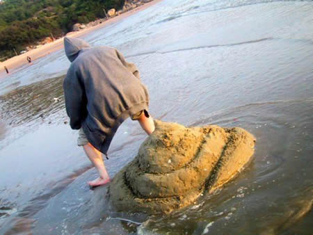 Person standing over giant pile of sand sculpted to look like a turd, so it looks like they pooped it out